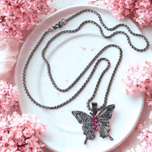 Collier Papillon Éternel argenté Envol Céleste posé sur assiette entourée de fleurs roses