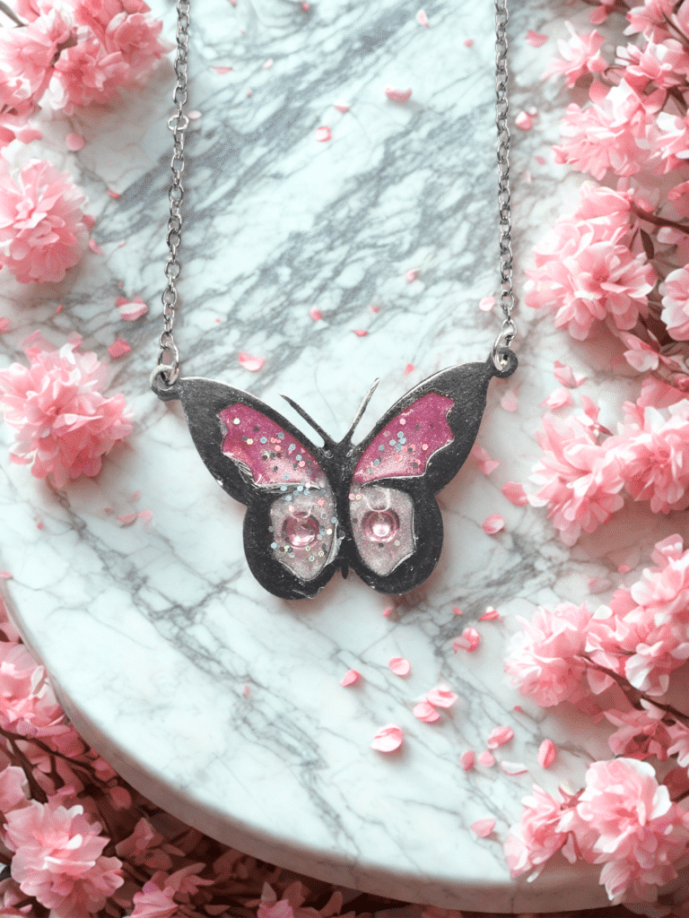 Collier Papillon Éternel rose pailleté Aile Enchantée finition argentée posé sur table